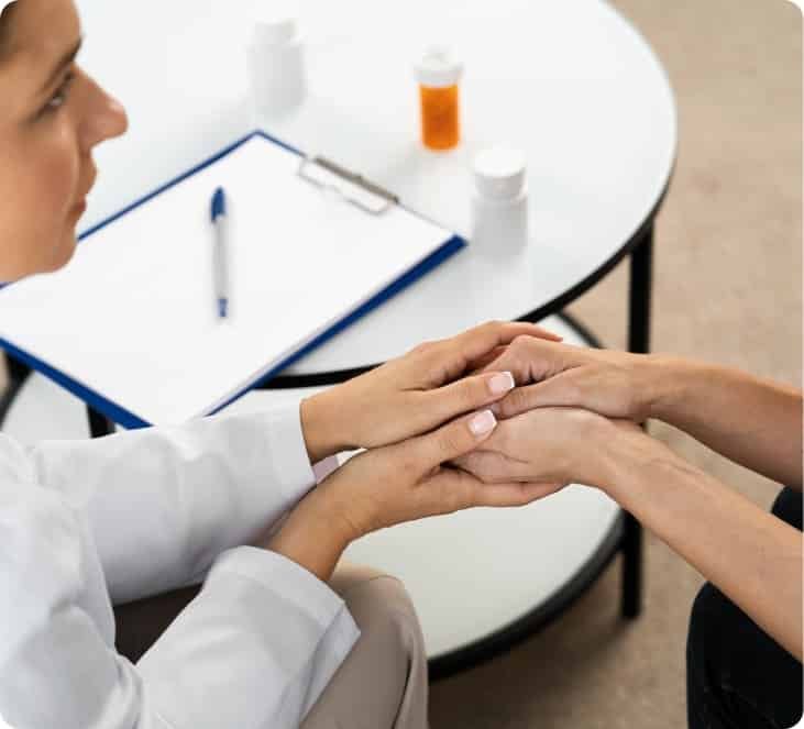 A healthcare professional holds a patient’s hands at a table with a clipboard, pen, and medication bottles in the background.