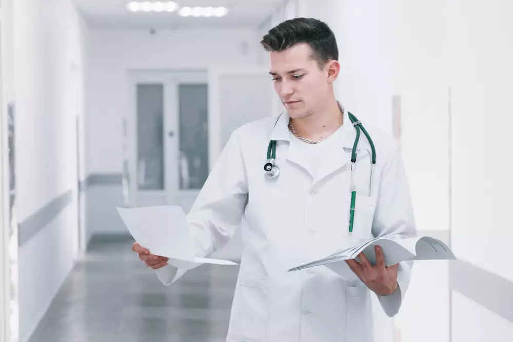 A doctor in a white coat holds paperwork and a medical book, standing in a bright hospital corridor.