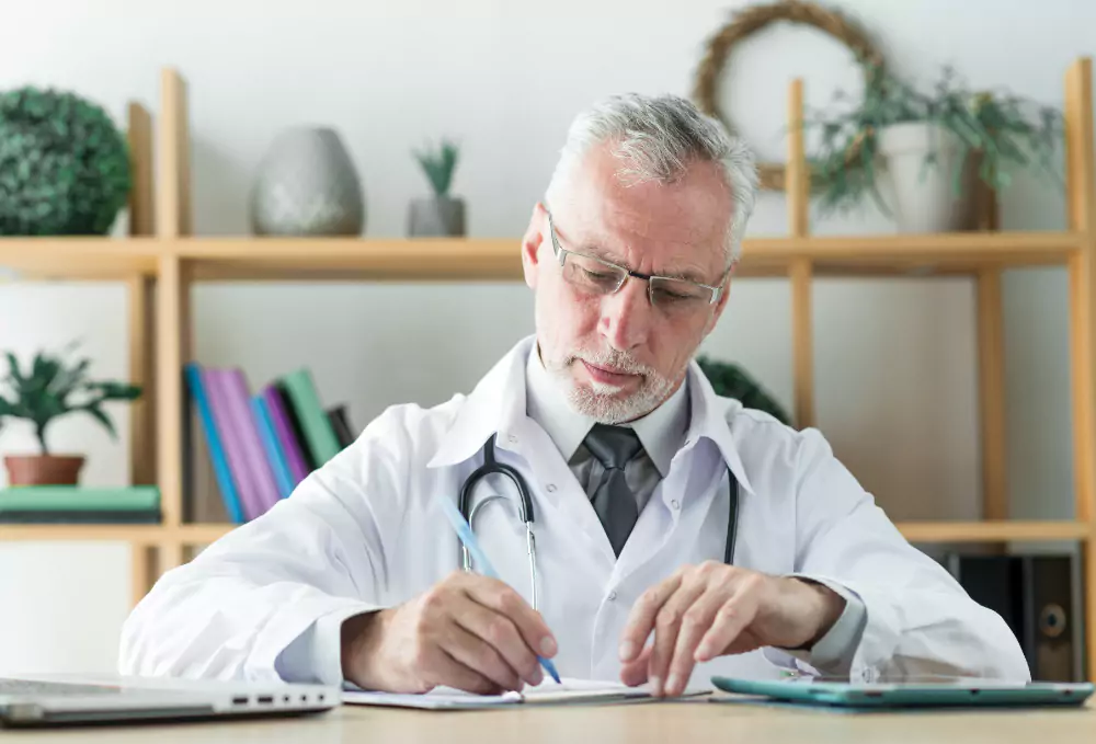 A medical professional in a white coat is writing notes at a desk, surrounded by plants and books on shelves.