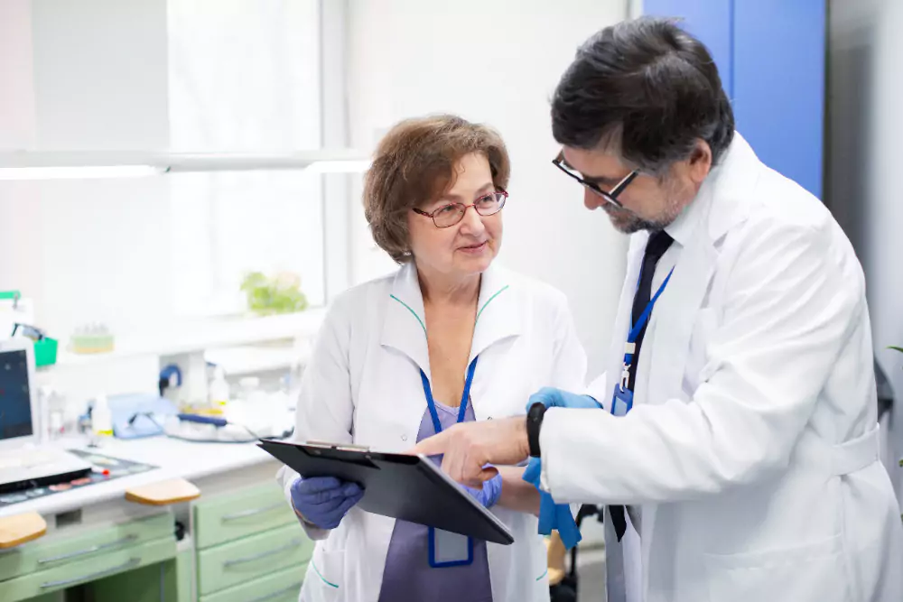 Two medical professionals in lab coats discuss patient information while reviewing a clipboard in a well-lit laboratory setting.