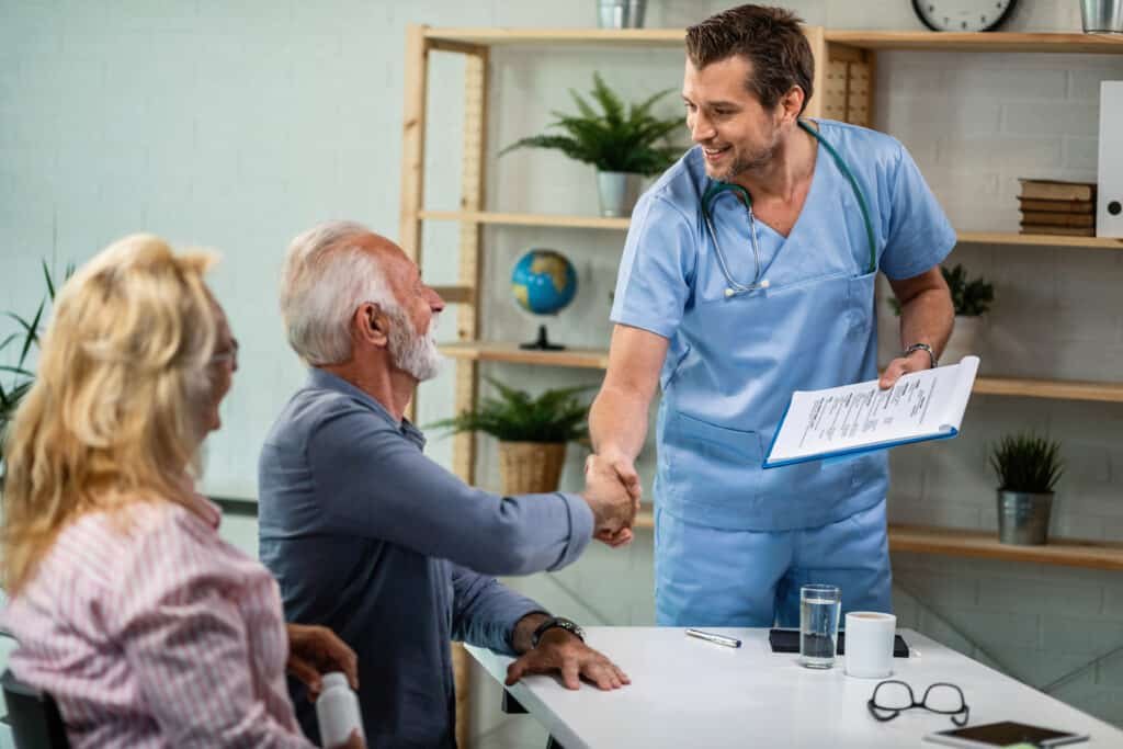 A smiling nurse in blue scrubs shakes hands with an elderly man at a table, with a woman nearby. The setting is a bright, welcoming clinic.
