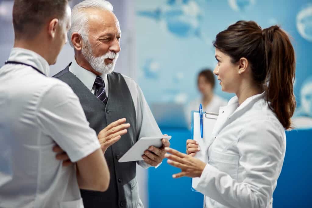 Three medical professionals engaged in discussion. An older man holds a tablet, while a woman holds a clipboard. The setting is a bright hospital corridor.