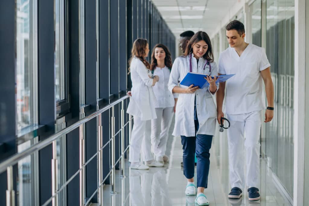 Young medical professionals in white coats walk down a bright hospital corridor. A woman reviews a chart with a man holding a stethoscope, radiating focus.