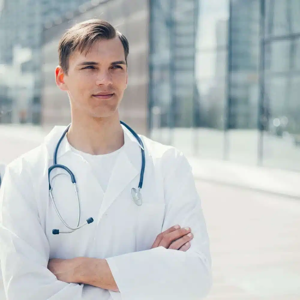 A male doctor poses in front of a healthcare facility, wearing a white coat and looking professional